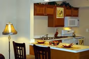 Interior view of a kitchen area with wooden cabinets, a white microwave, stove, and countertop. The countertop has place settings with plates and bowls, and there are two wooden chairs in front of it. A decorative lamp is visible on the left side.