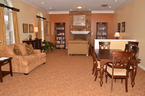 A cozy living room area with beige carpet and walls painted in warm tones. The room features a tan sofa with patterned cushions, a wooden dining table with six chairs, two tall bookshelves flanking a stone fireplace with a mantle, framed artwork on the walls, and windows with beige curtains allowing natural light to enter.
