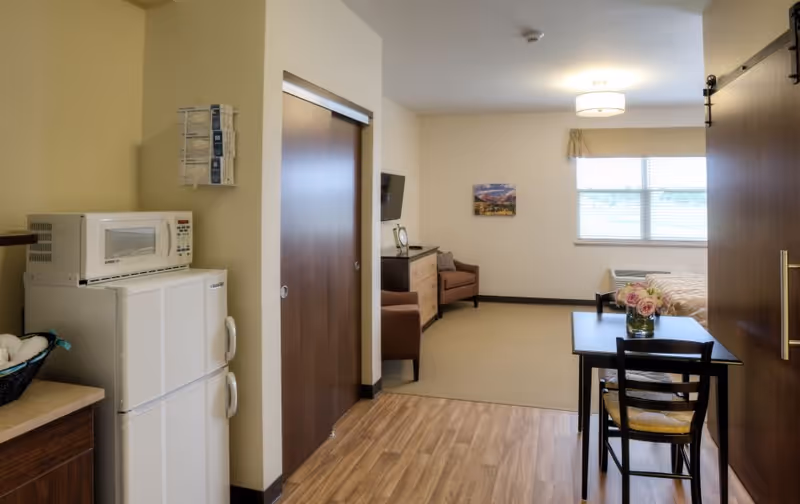 Interior view of a senior living facility room showing a small kitchenette area with a microwave and refrigerator on the left, a dining table with two chairs and a vase of flowers in the foreground, and a living area with an armchair, dresser, wall-mounted TV, and a window with blinds in the background.