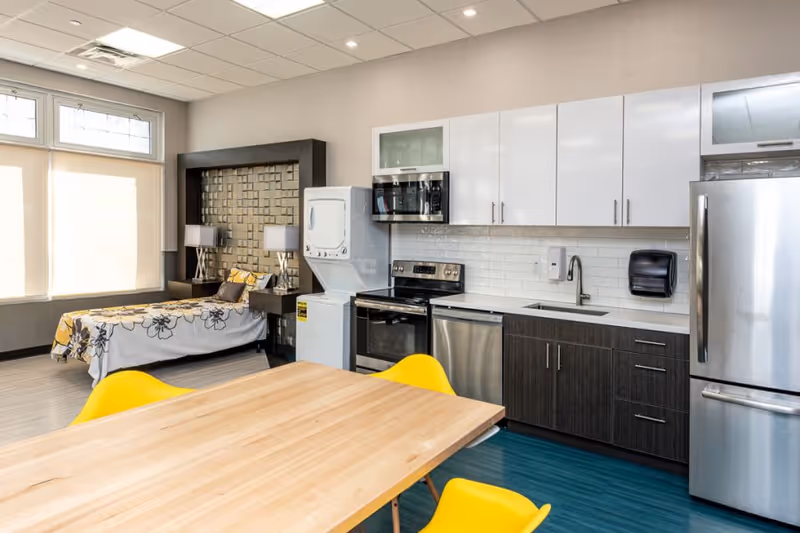 A modern studio apartment-style room with a kitchen and sleeping area. The kitchen features white upper cabinets, dark lower cabinets, a stainless steel refrigerator, dishwasher, oven, microwave, and a stacked washer and dryer. A wooden dining table with bright yellow chairs is in the foreground. The sleeping area has a bed with a floral bedspread and two bedside tables with lamps. Large windows with blinds allow natural light into the room.
