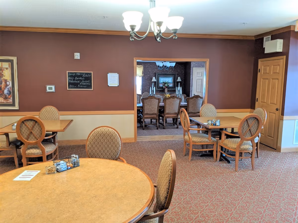 Interior view of a dining area in a senior living facility named Deer Crest, featuring round and square tables with patterned upholstered chairs, a chandelier hanging from the ceiling, a brown and beige color scheme, and an adjoining room with a larger dining table and chairs visible through an open doorway.