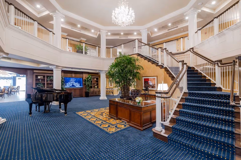 Spacious and elegant senior living facility lobby with a grand staircase covered in blue carpet with gold dots, white railings, a large chandelier hanging from the ceiling, a black grand piano on the left, a wooden desk with a plant and lamp in the center, and a large green plant behind the desk. The area has blue carpet flooring with a patterned rug under the desk, white columns, and a second-floor balcony overlooking the lobby.