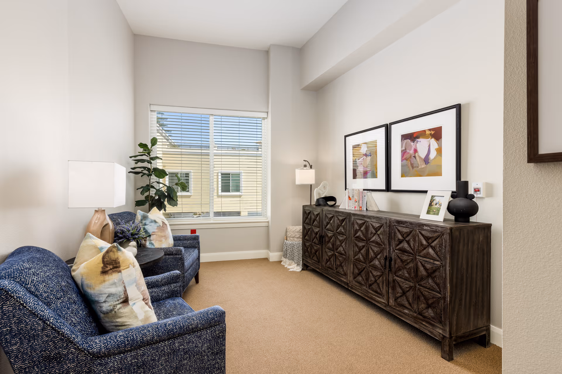 A cozy sitting area with two blue upholstered armchairs adorned with decorative pillows, a small round table between them holding a plant, a large window with blinds letting in natural light, and a dark wooden sideboard against the opposite wall displaying framed artwork, books, and decorative items.