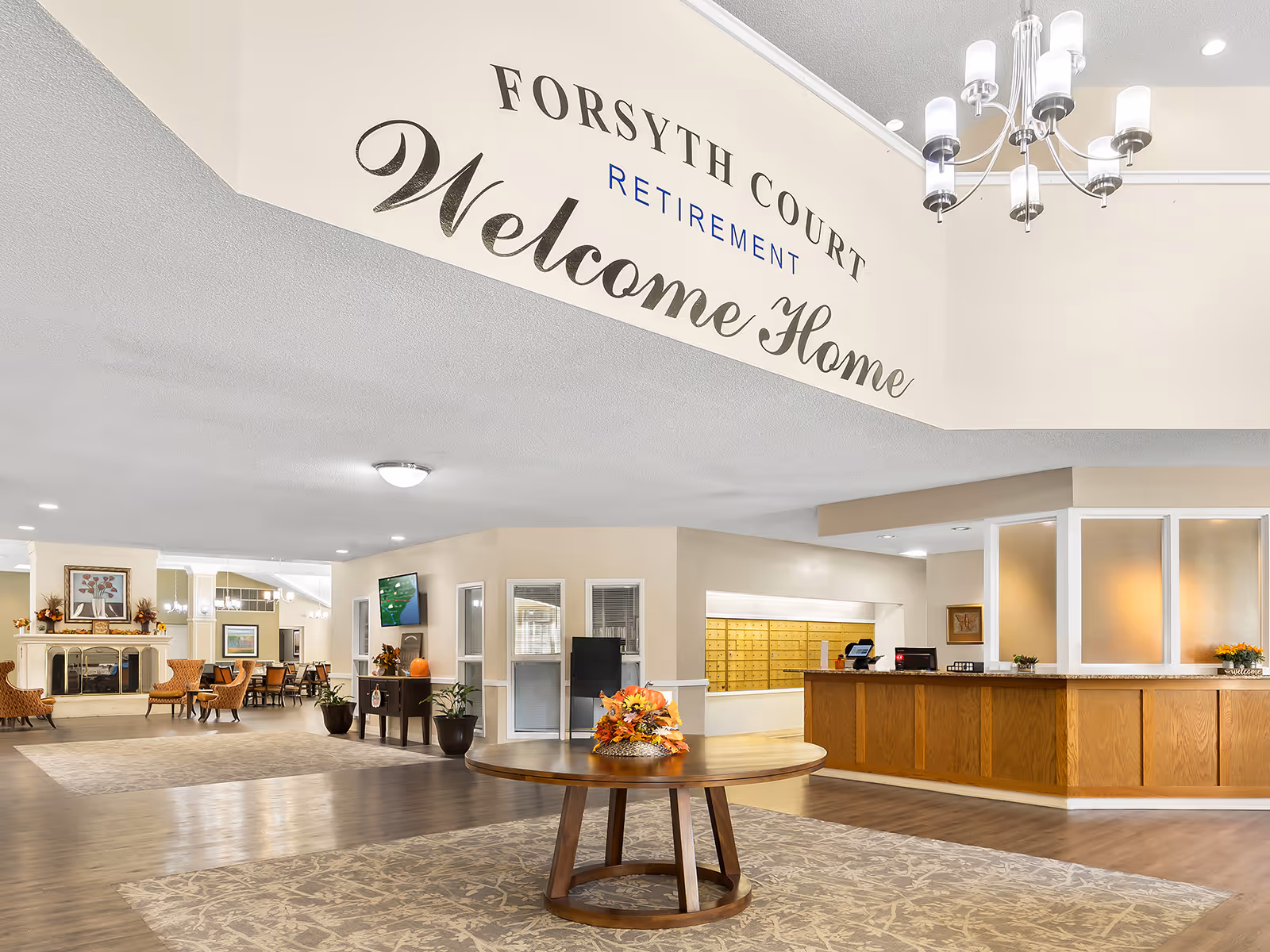 Interior view of the Holiday Forsyth Court retirement facility lobby featuring a round wooden table with a floral centerpiece, a reception desk with frosted glass panels, a seating area with chairs and a fireplace, and a large wall sign that reads 'Forsyth Court Retirement Welcome Home'.