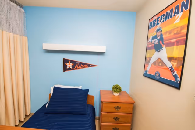 A small bedroom with a single bed featuring a navy blue bedspread and pillow. Above the bed on a light blue wall is a Houston Astros pennant. Next to the bed is a wooden nightstand with three drawers and a small potted plant on top. On the adjacent beige wall is a framed poster of a baseball player in a batting stance.
