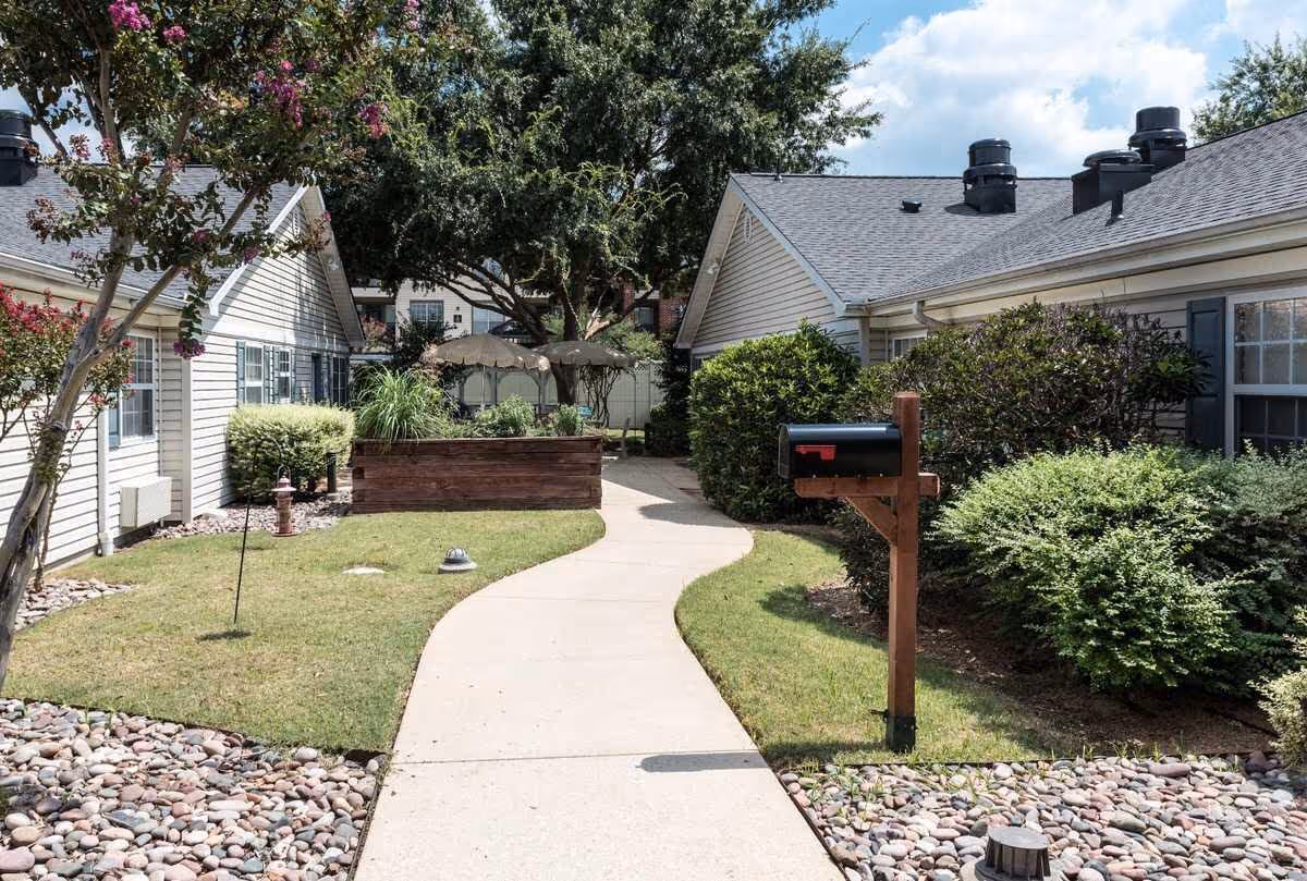 A paved walkway curves through a landscaped outdoor area between two single-story buildings with beige siding and dark shutters. There are bushes, trees, and a mailbox on a wooden post along the path. In the background, there are patio umbrellas and more greenery under a partly cloudy sky.