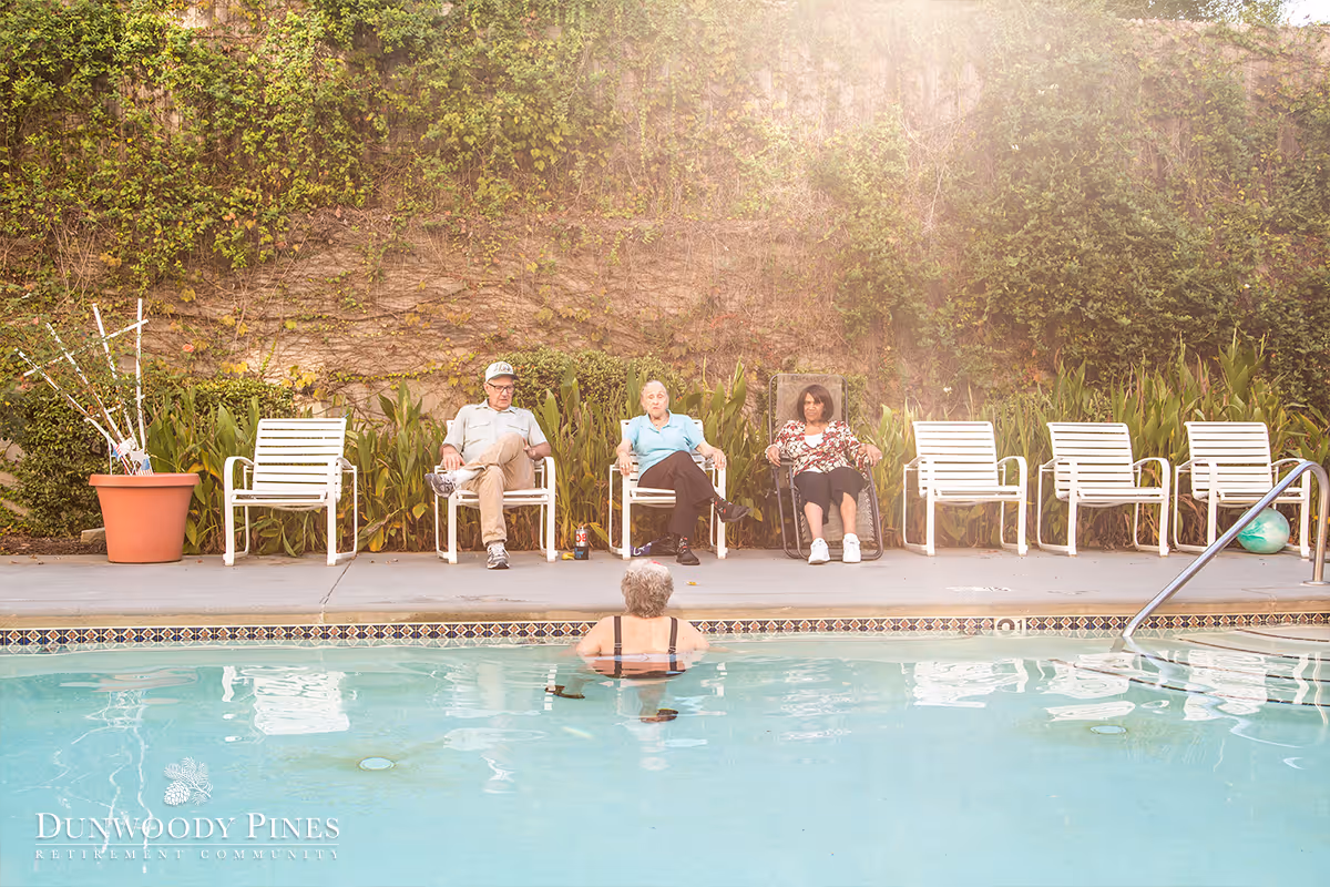 Three elderly people sitting on white chairs by the side of an outdoor swimming pool, with one person in the pool. The background shows a wall covered with greenery and plants, and there is a large potted plant on the left side.