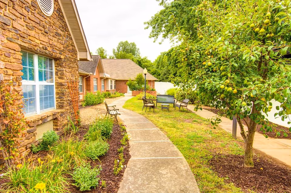 A paved walkway curves through a garden area with green grass, plants, and a tree bearing fruit. On the left side, there is a building with stone and brick walls and windows. Along the path, there are several benches and chairs arranged for seating. The scene is bright and appears to be outdoors in a peaceful setting.