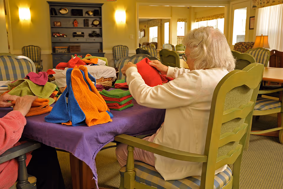An elderly woman sitting at a table folding colorful towels in a well-lit room with green chairs and a purple tablecloth. Another person is partially visible folding towels on the left side. The room has a cozy atmosphere with soft lighting and a cabinet with decorative items in the background.
