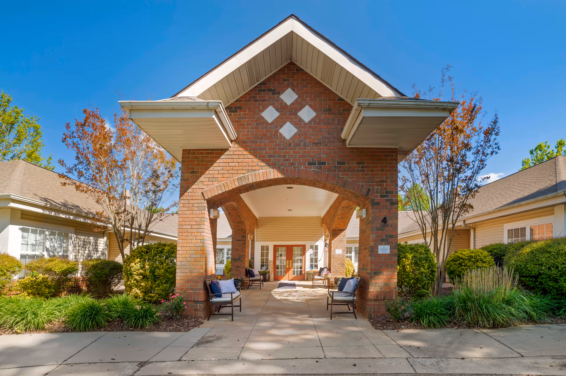 Entrance of a senior living facility with a brick archway and covered walkway. There are benches with cushions on either side of the walkway, surrounded by well-maintained bushes and trees under a clear blue sky.