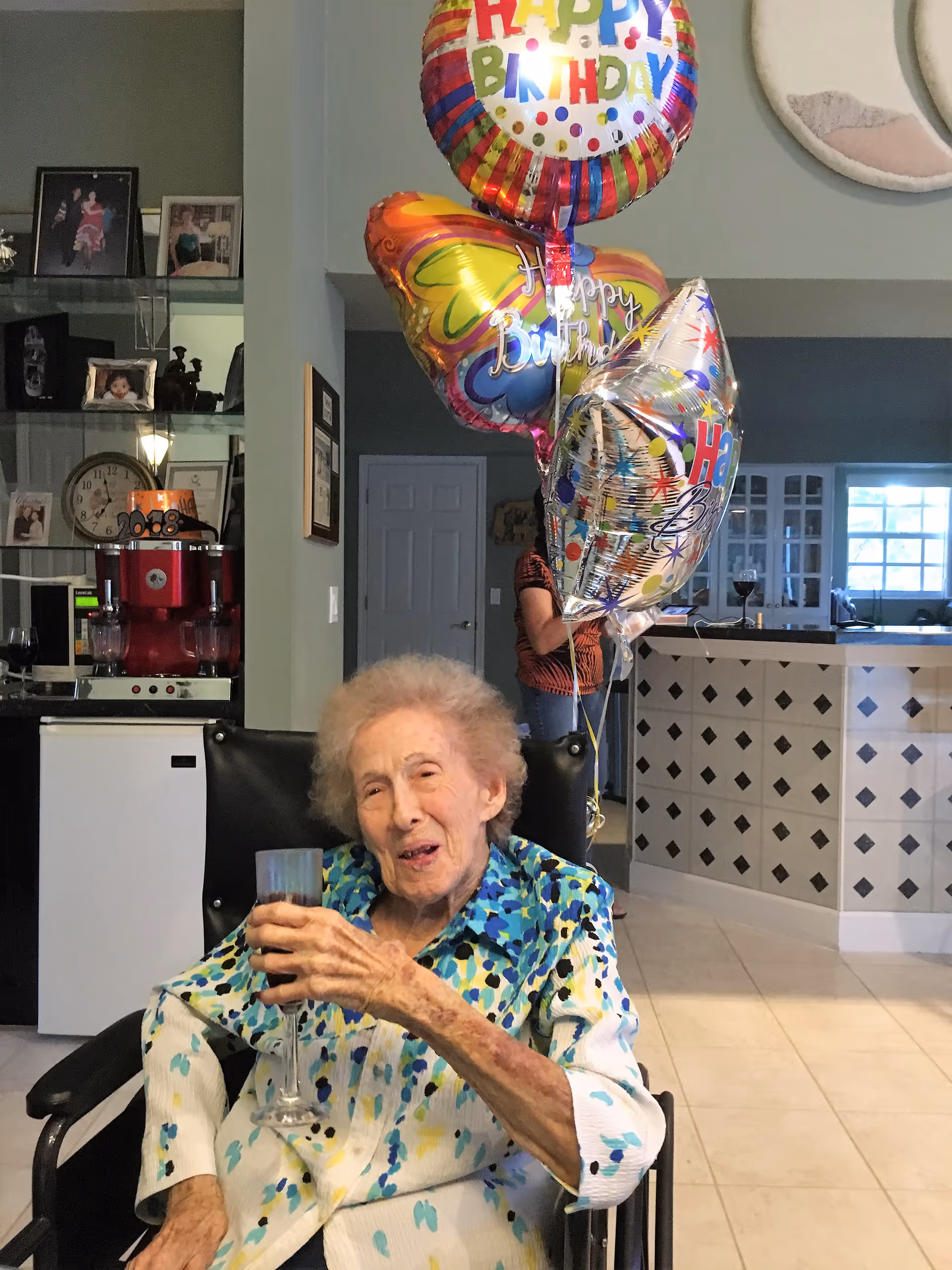 An elderly woman sitting in a wheelchair holding a glass, smiling. Behind her are colorful 'Happy Birthday' balloons. The setting appears to be a kitchen or dining area with cabinets, a counter, and a person partially visible in the background.