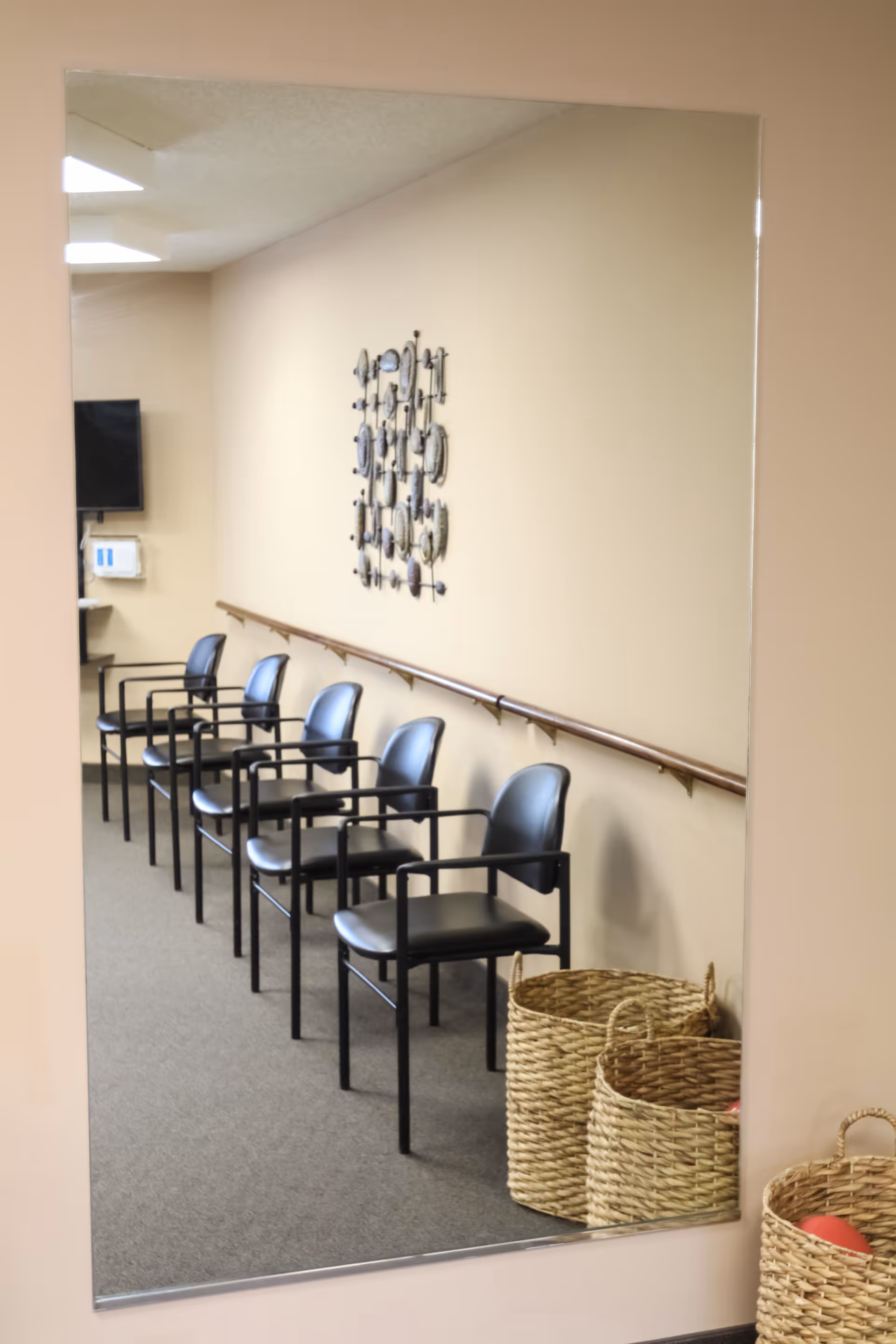 A reflection in a large wall mirror showing a row of black chairs with armrests lined up against a beige wall. Above the chairs is a decorative wall art piece. There is a wooden handrail running along the wall behind the chairs. On the floor near the mirror are three woven baskets, one containing a red ball. The room has a carpeted floor and a ceiling with fluorescent lights.