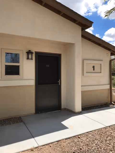 Exterior view of a beige building with a dark door and two windows. The building has a number '1' displayed on the wall to the right of the door. There is a small light fixture above the door and a concrete walkway in front of the entrance. The sky is partly cloudy.
