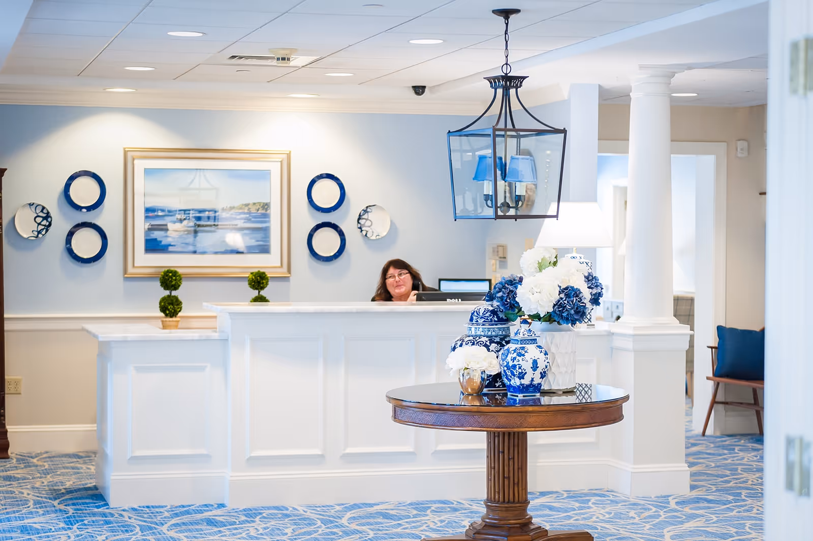 Bright reception area with a white front desk, a staff member seated behind it, and a round table displaying blue-and-white vases and flowers.