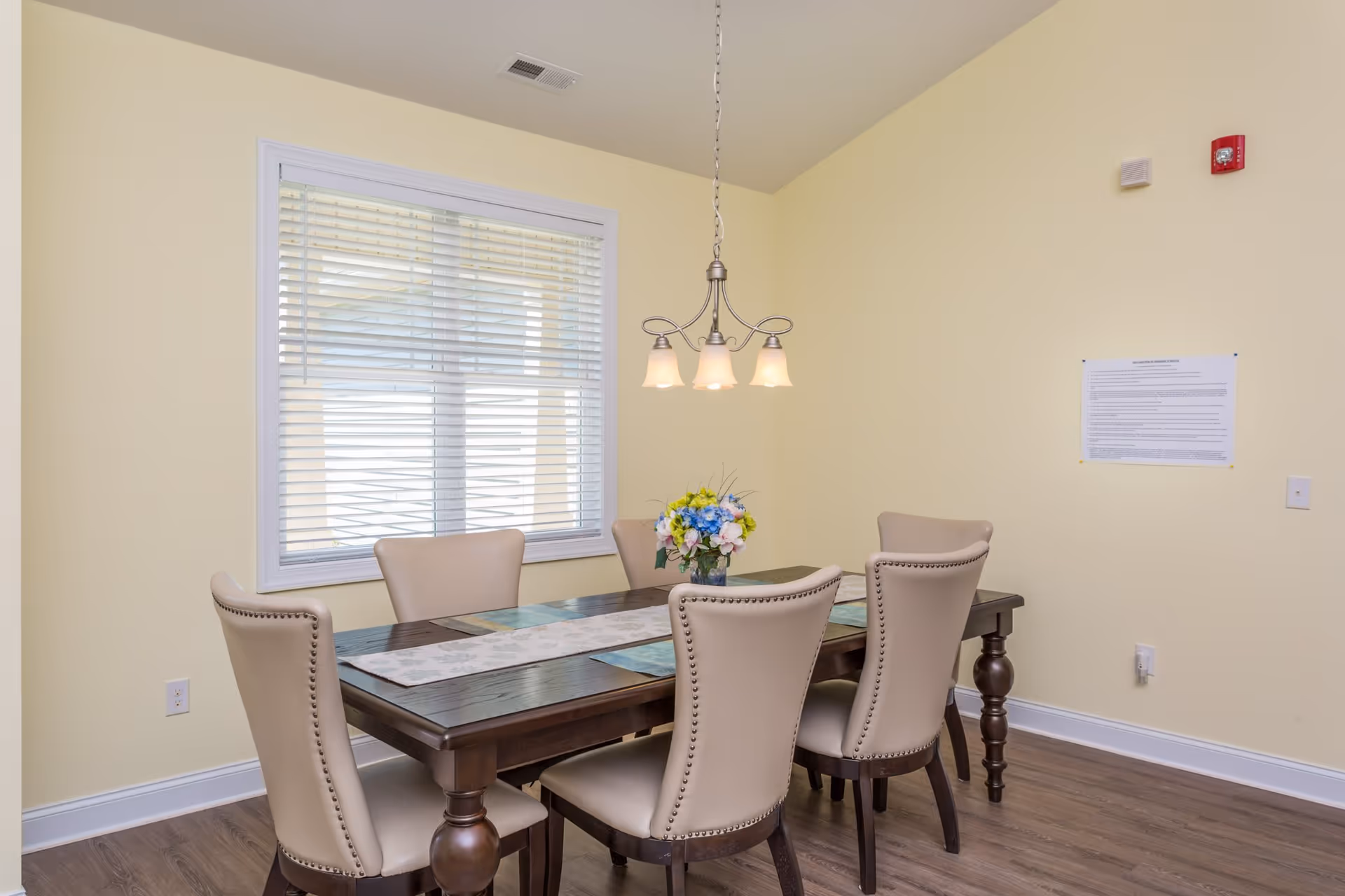 A dining room with a dark wooden table surrounded by six beige upholstered chairs. A floral centerpiece is placed on the table along with placemats and a table runner. A window with white blinds is on the left wall, and a three-light chandelier hangs above the table. The walls are painted light yellow and the floor is wood.