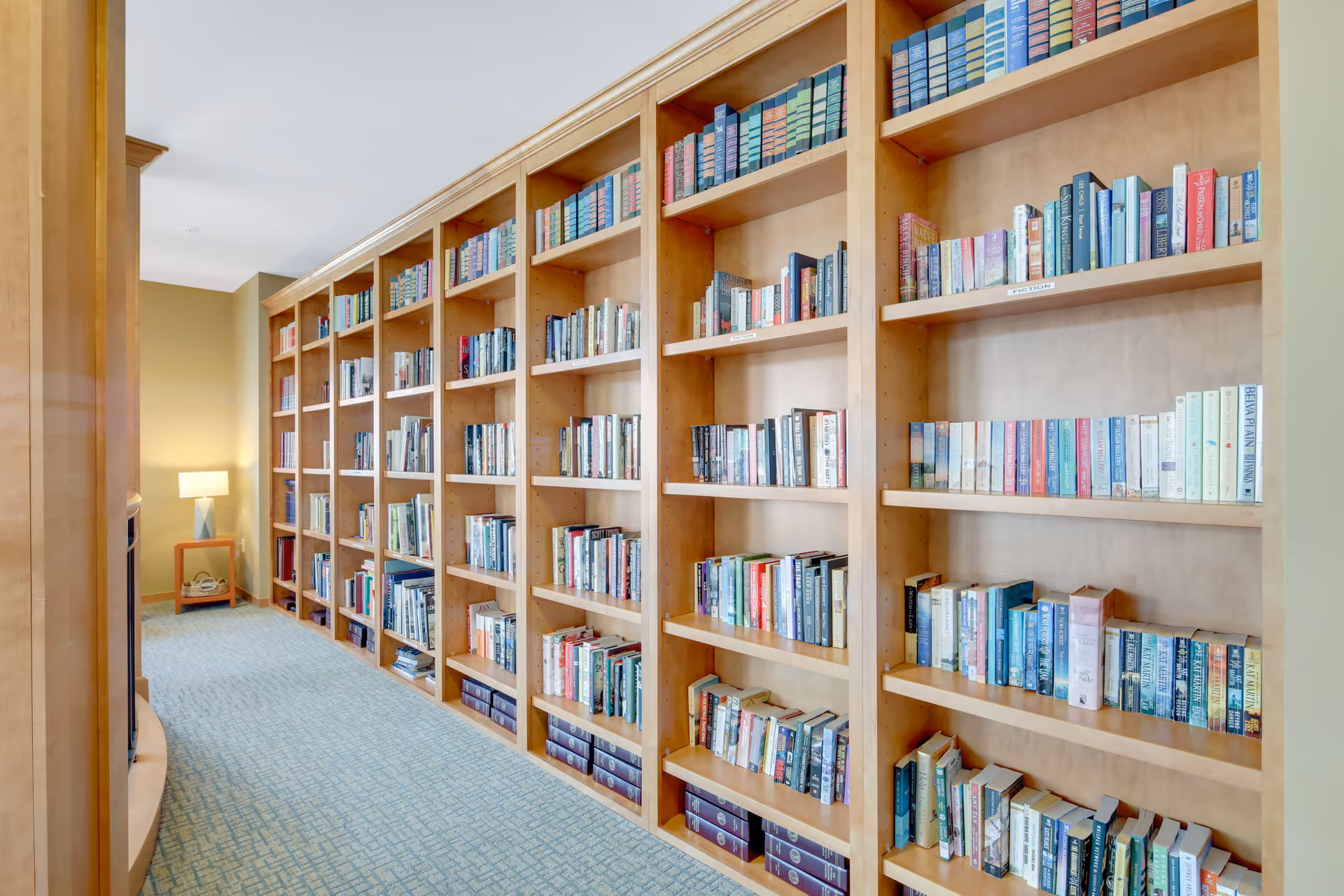 A well-lit hallway lined with tall wooden bookshelves filled with a variety of books. At the end of the hallway, there is a small wooden table with a modern lamp on it, and the walls are painted in a warm beige color. The floor is carpeted with a patterned blue carpet.