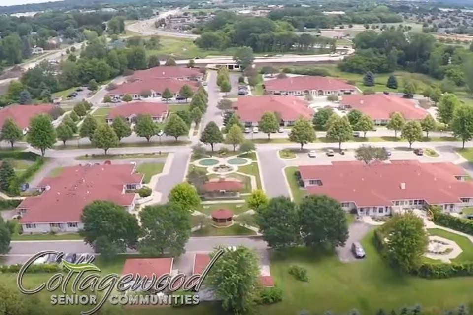 Aerial view of Cottagewood Senior Communities showing multiple single-story buildings with red roofs arranged around landscaped gardens and tree-lined pathways. There is a central circular garden area with a gazebo and decorative landscaping.