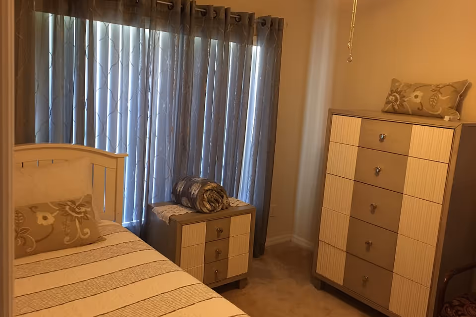 A cozy bedroom with a single bed featuring a striped bedspread and a decorative pillow. Next to the bed is a small nightstand with a rolled blanket on top. A tall dresser with multiple drawers and a matching decorative pillow on top stands against the wall. The room has beige walls and carpet, with a window covered by blue curtains.