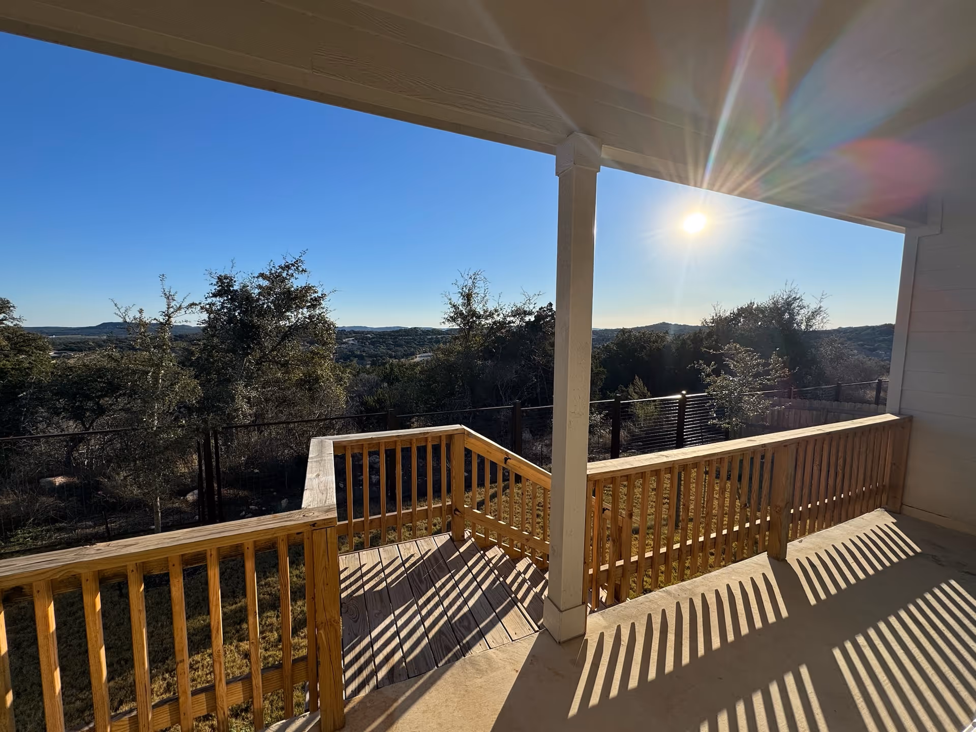 View from a covered wooden porch overlooking a fenced backyard with trees and hills in the distance under a clear blue sky with the sun shining brightly.