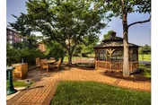 Sunny outdoor courtyard with a wooden gazebo, patio dining set, brick pathways, and trees.