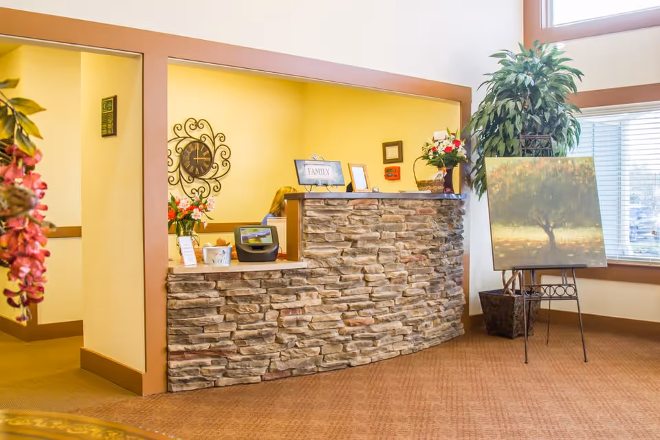 Reception area with a stone facade desk, a person working behind the desk, a decorative wall clock, flowers, a framed sign that says FAMILY, and a large potted plant next to an easel holding a painting of a tree near a window with blinds.