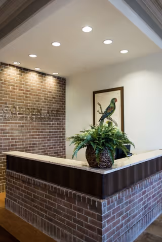 Reception desk in a senior living facility lobby with exposed brick walls, a marble countertop, a potted fern, and framed bird artwork.