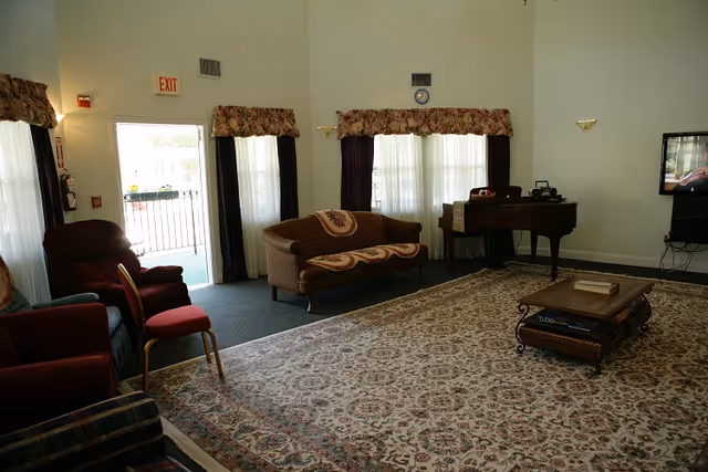 A spacious living room with a large patterned area rug covering most of the floor. There are several upholstered chairs and a sofa with decorative cushions. A grand piano is positioned against the far wall near windows with floral valances and sheer curtains. A flat-screen TV is mounted on the right wall. The room has light-colored walls and an exit door with an illuminated exit sign above it.