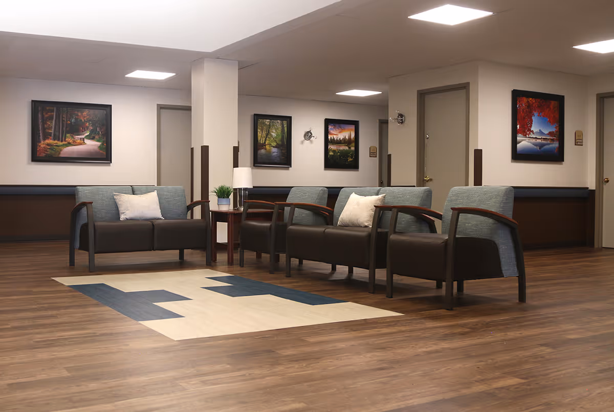 A nursing home lounge with sofas and armchairs arranged around a rug and side tables beneath framed artwork.