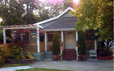 Front exterior view of a small residential-style building with a porch, two potted evergreen plants flanking the entrance steps, a white chair on the right side, and a wooden fence surrounding the property. The building number 9644 is displayed vertically on a porch column. Trees and greenery surround the building.