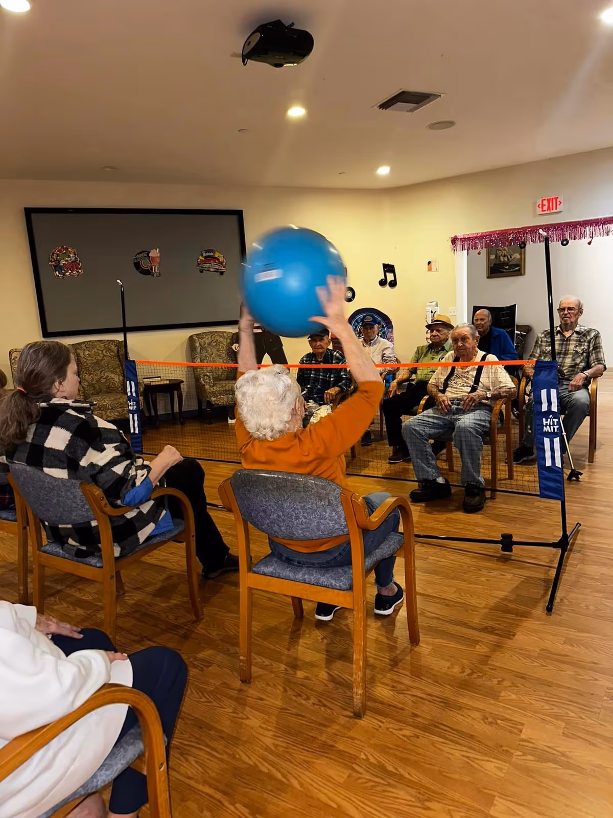 A group of elderly people seated in chairs arranged in two rows facing each other in a room with wooden flooring. They are playing a seated volleyball game with a blue exercise ball over a small net. The room has beige walls with some decorations and an exit sign above a door in the background.