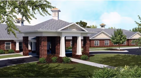 Exterior view of a single-story brick building with a covered entrance supported by white columns, surrounded by green grass, shrubs, and trees under a blue sky with some clouds.