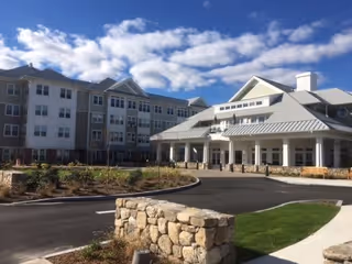 Exterior view of a large, multi-story senior living facility with a covered entrance, stone accents, and a circular driveway under a partly cloudy sky.