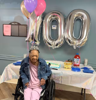 An elderly woman in a wheelchair wearing a tiara and a denim jacket, sitting in front of a table decorated for a celebration with large silver balloons forming the number 100, colorful balloons, a cake, and party supplies in a room with a menu board on the wall.