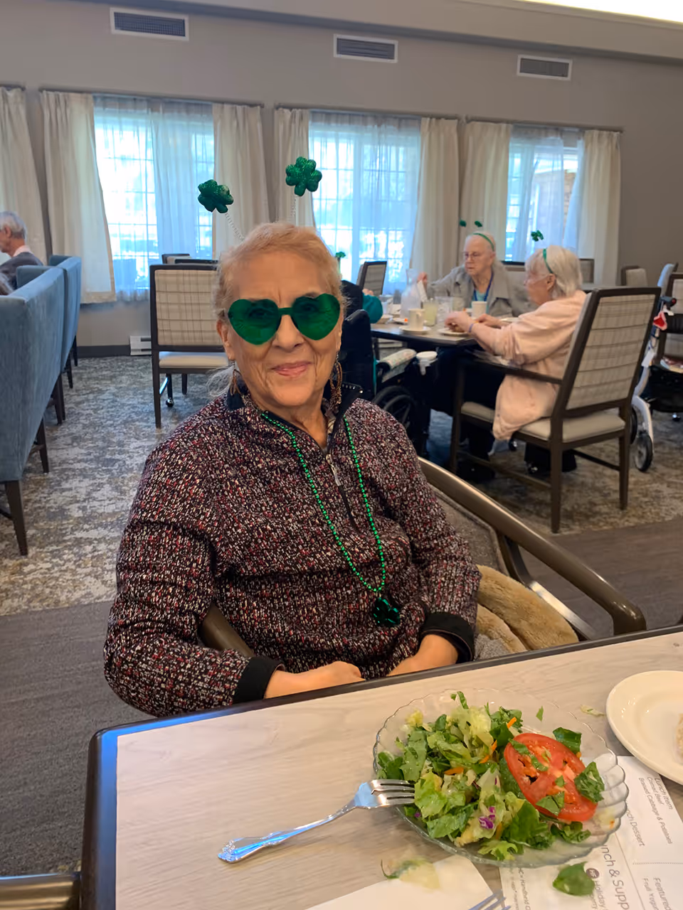 An elderly woman wearing green shamrock glasses and beads sits at a dining table with a salad while other residents eat in the background.