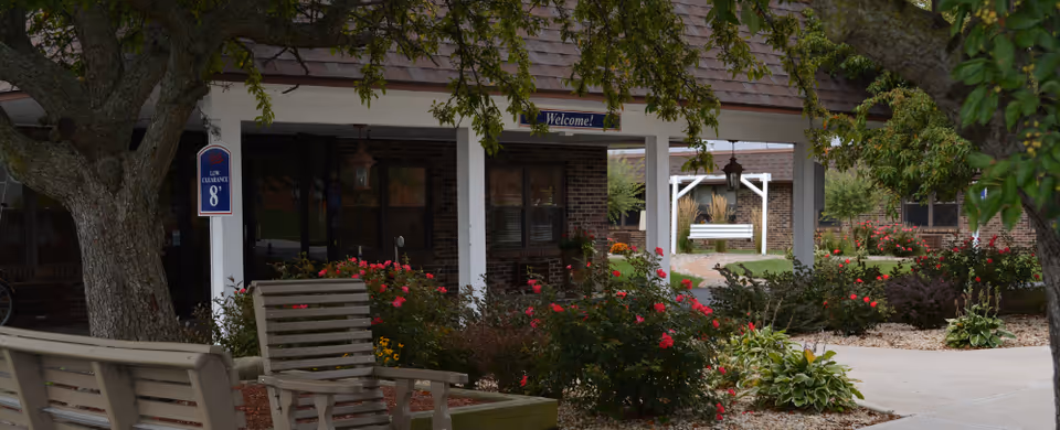 Outdoor garden area at Valparaiso Care and Rehabilitation featuring wooden benches, blooming rose bushes, trees, and a covered porch with a 'Welcome!' sign.