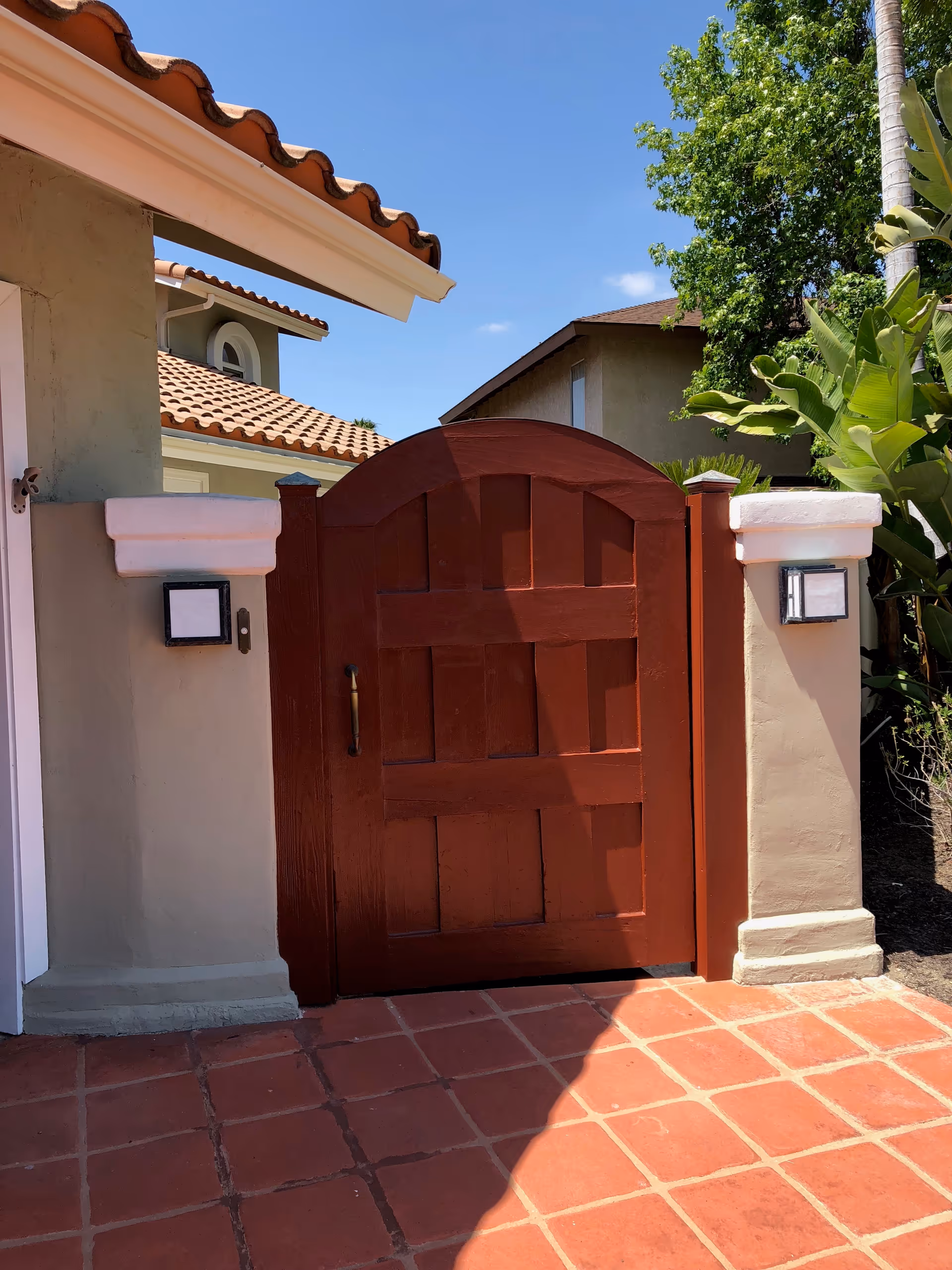 Red wooden arched gate set between stucco pillars with terracotta tile roof and a tiled patio, with plants and neighboring houses visible.