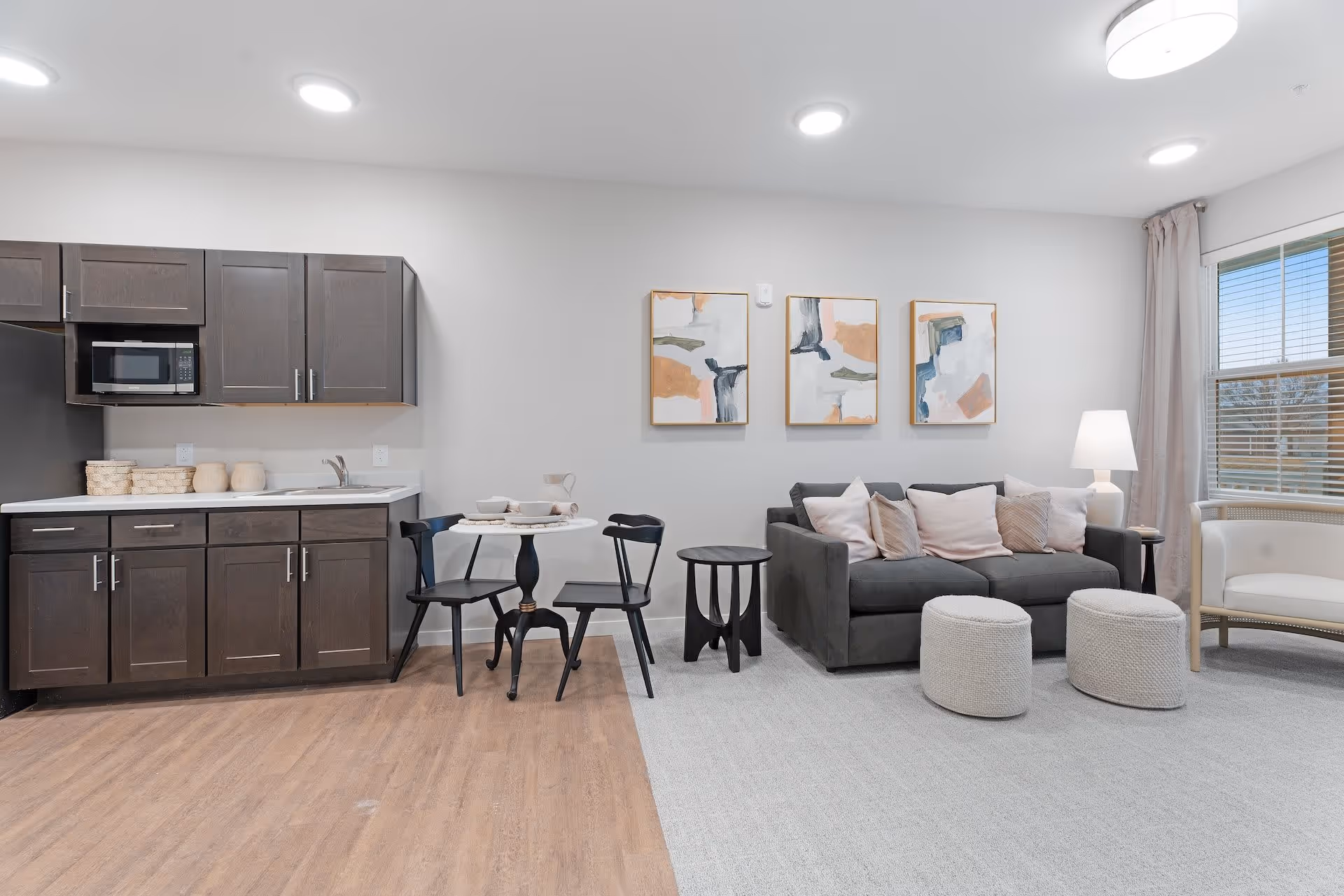 A modern senior living space featuring a small kitchen area with dark wood cabinets, a microwave, and a sink. Adjacent to the kitchen is a small round dining table with two black chairs. The living area includes a gray sofa with multiple light-colored pillows, two round knitted ottomans, a side table, a lamp, and a white armchair near a window with blinds and light curtains. Three abstract paintings hang on the wall above the sofa.