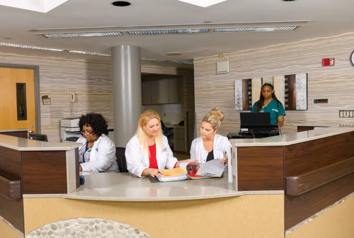 A reception or nurse station area inside Elmwood Hills Healthcare Center with three healthcare workers. Two women in white lab coats are seated at the counter reviewing documents, while another woman in green scrubs is standing behind a computer. The area has a modern design with a curved counter, wood paneling, and textured wallpaper.
