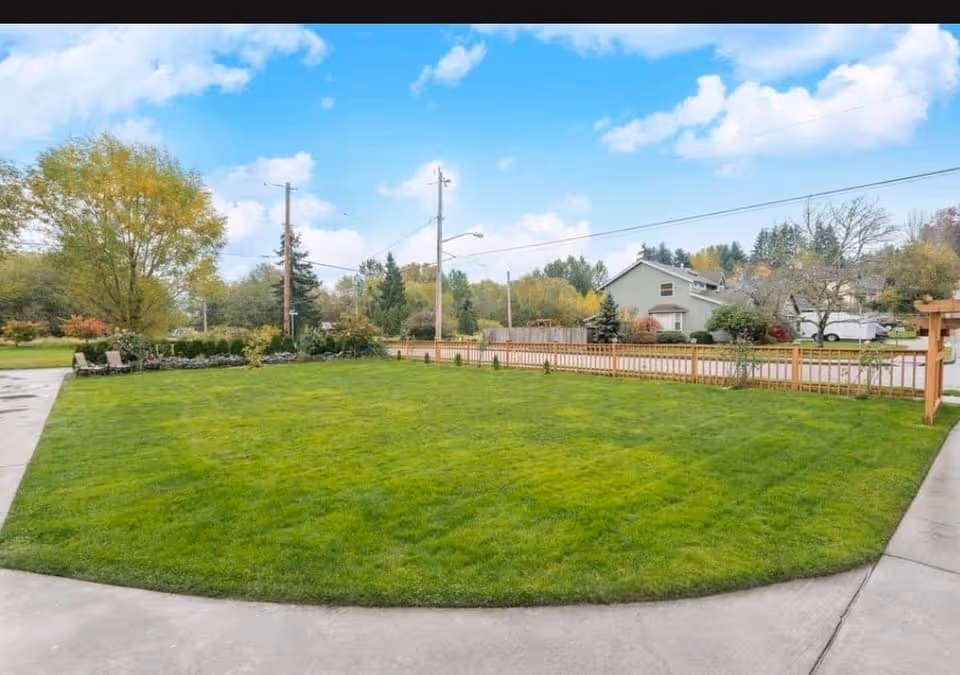 A well-maintained green lawn bordered by a wooden fence with a few small plants along the fence line. There are trees and houses in the background under a partly cloudy blue sky. A concrete pathway curves around the lawn area.