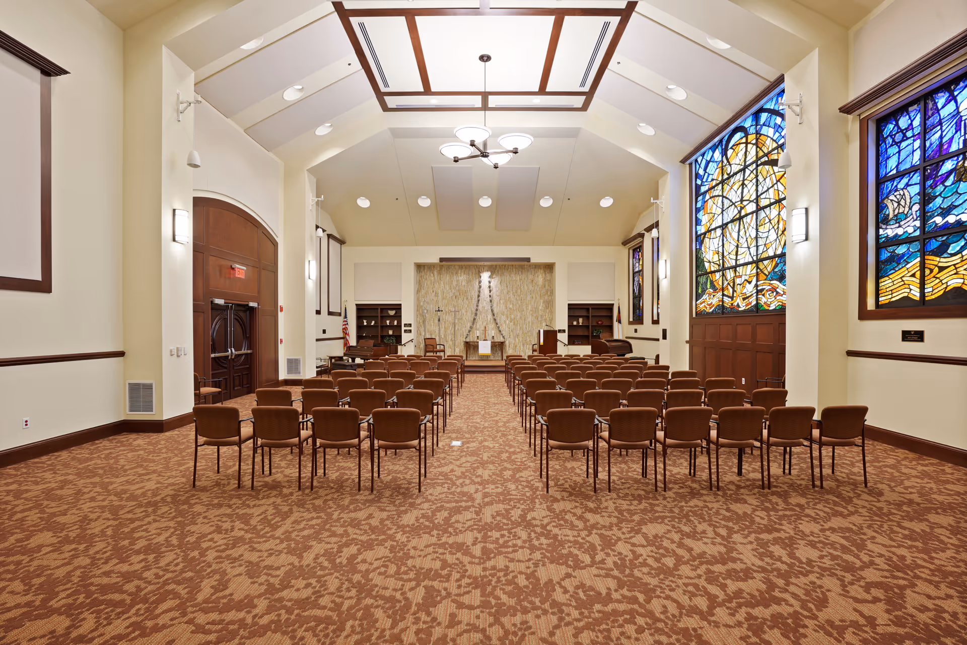 A large, well-lit chapel or auditorium with rows of brown chairs facing a stage area with a podium and a cross on the back wall. The room features high ceilings with recessed lighting and a chandelier, large stained glass windows on the right side, and wood trim accents throughout.
