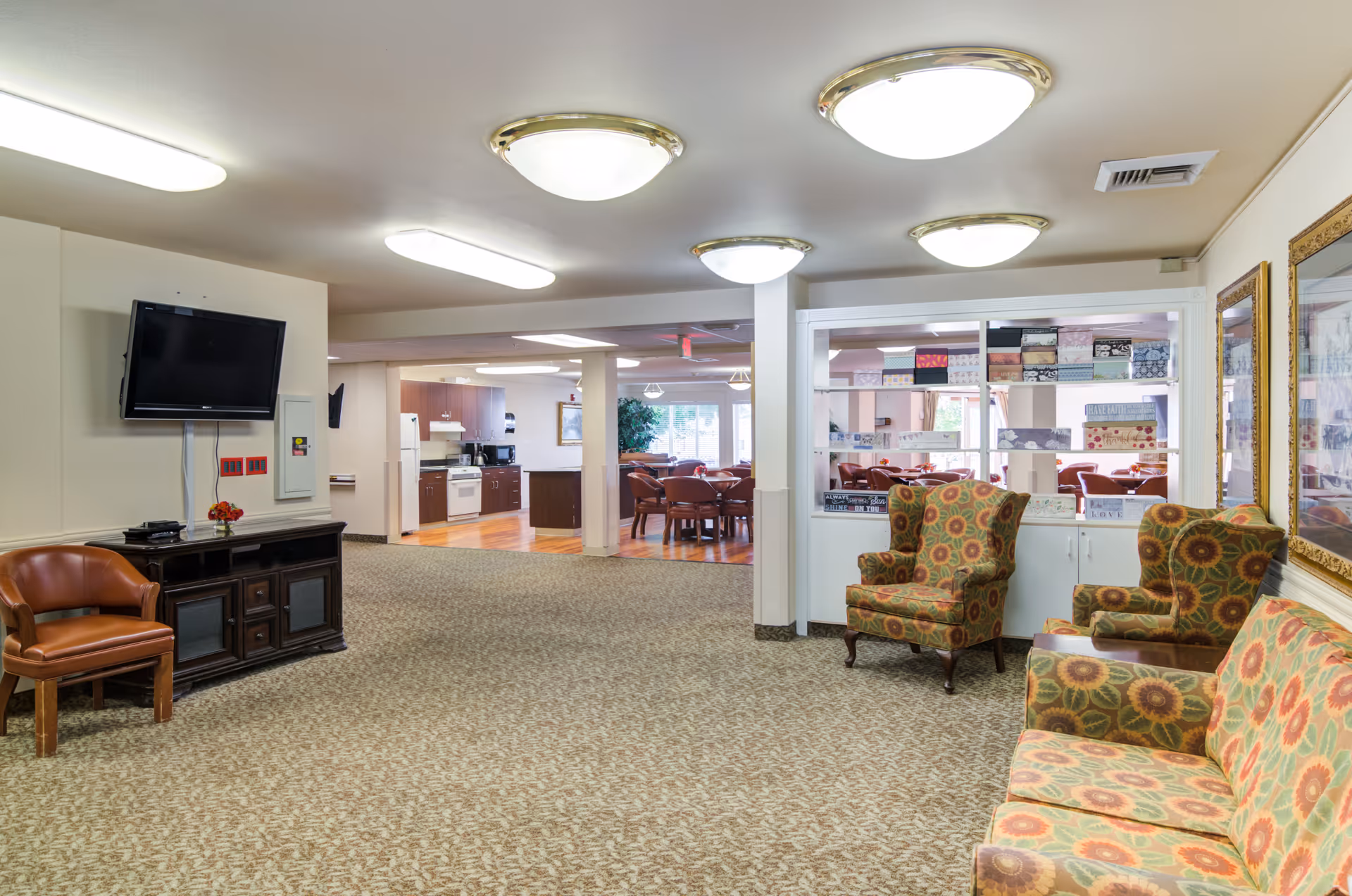 Interior view of a senior living facility common area with patterned armchairs and a sofa on the right, a wall-mounted TV on the left, and a kitchen and dining area visible in the background.