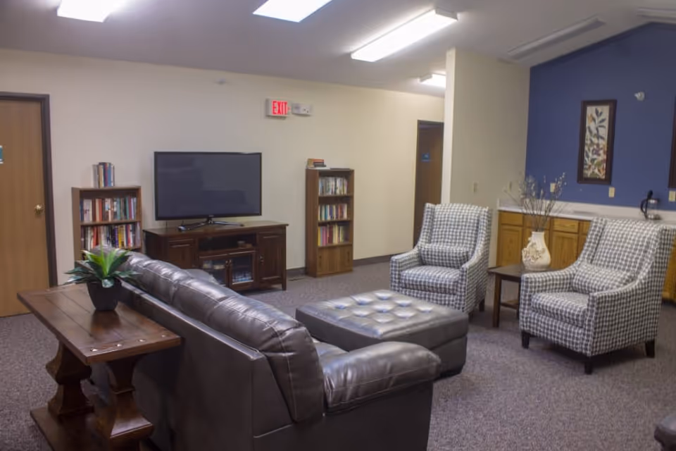 A cozy living room area with a dark brown leather sofa, two patterned armchairs, and a tufted ottoman in the center. There is a wooden side table with a potted plant behind the sofa, a TV on a wooden stand, and two bookshelves filled with books against the wall. The walls are light-colored with one accent wall painted blue, decorated with framed artwork and a vase with dried branches on a small table.