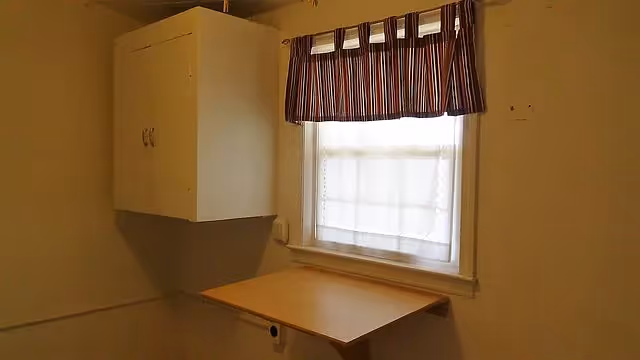 Small interior room with a window covered by a striped valance, a wall-mounted cabinet, and a fold-down wooden table.