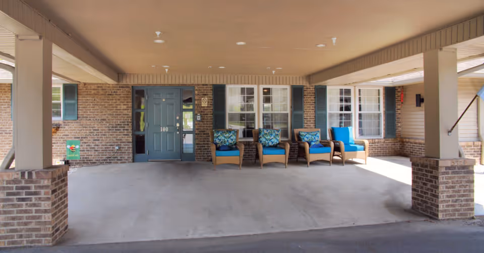 Covered entrance area of a building with a blue door labeled 100, brick walls, and four wicker chairs with blue cushions and patterned pillows arranged in a row near the windows.