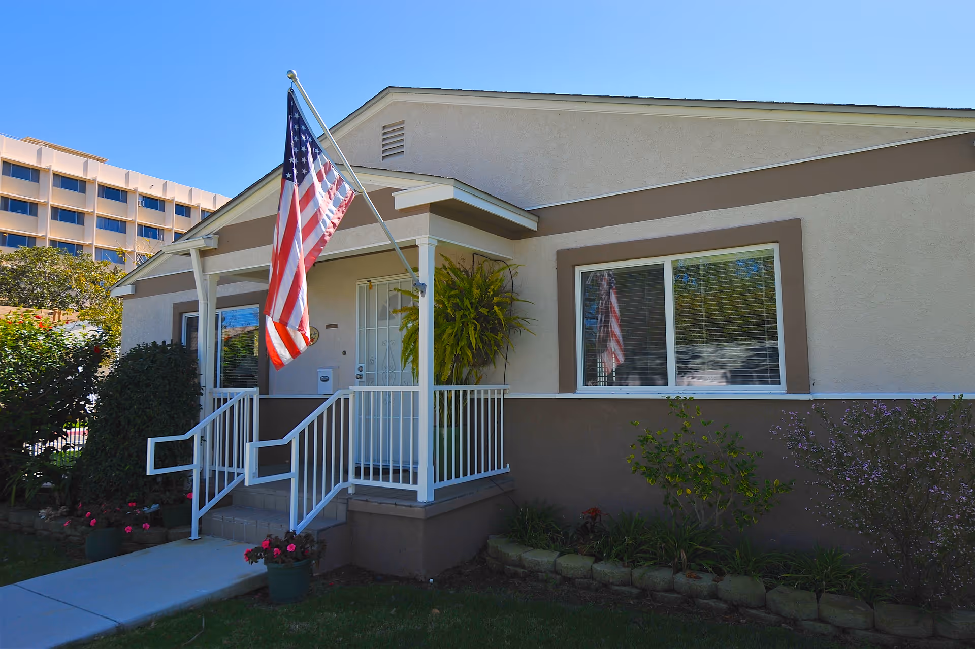 Exterior view of a single-story building with beige and brown walls, a small porch with white railings, an American flag mounted on a pole, and some plants and bushes around the entrance. A multi-story building is visible in the background under a clear blue sky.