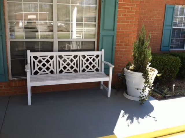 White wooden bench and large potted plant on a porch in front of a brick building with green shutters and a window.