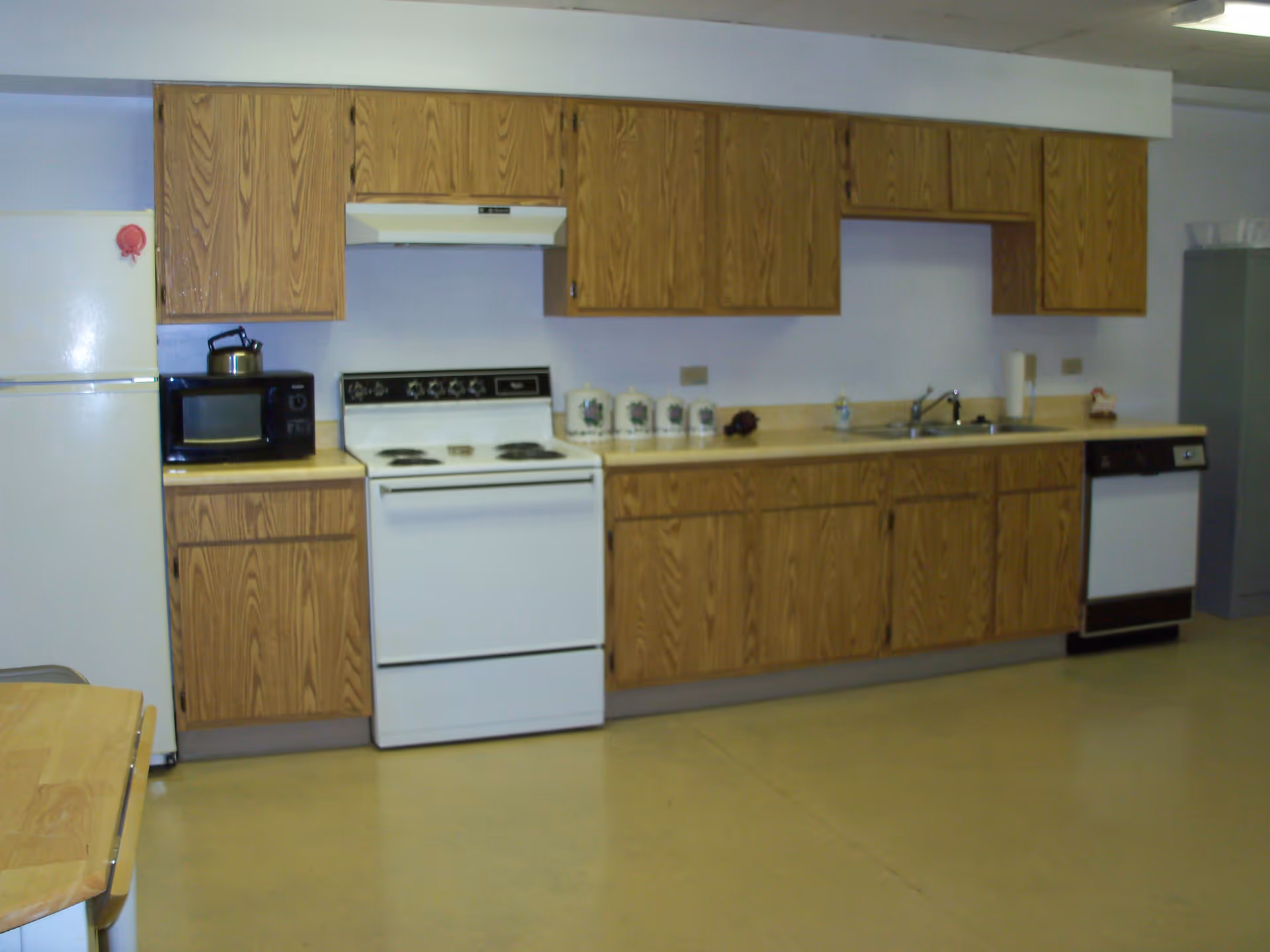 A kitchen with wooden cabinets, a white refrigerator, a microwave, a white electric stove with an overhead vent, a countertop with canisters, a sink, and a dishwasher. The floor is a light beige color and the walls are light-colored.