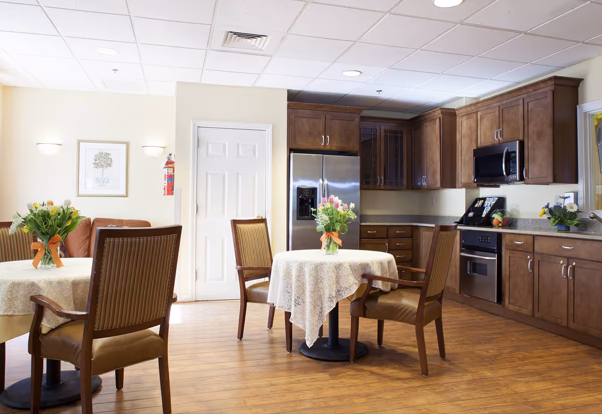 Well-lit communal dining area with round tables and chairs in front of a full kitchen with wooden cabinets and stainless steel appliances.