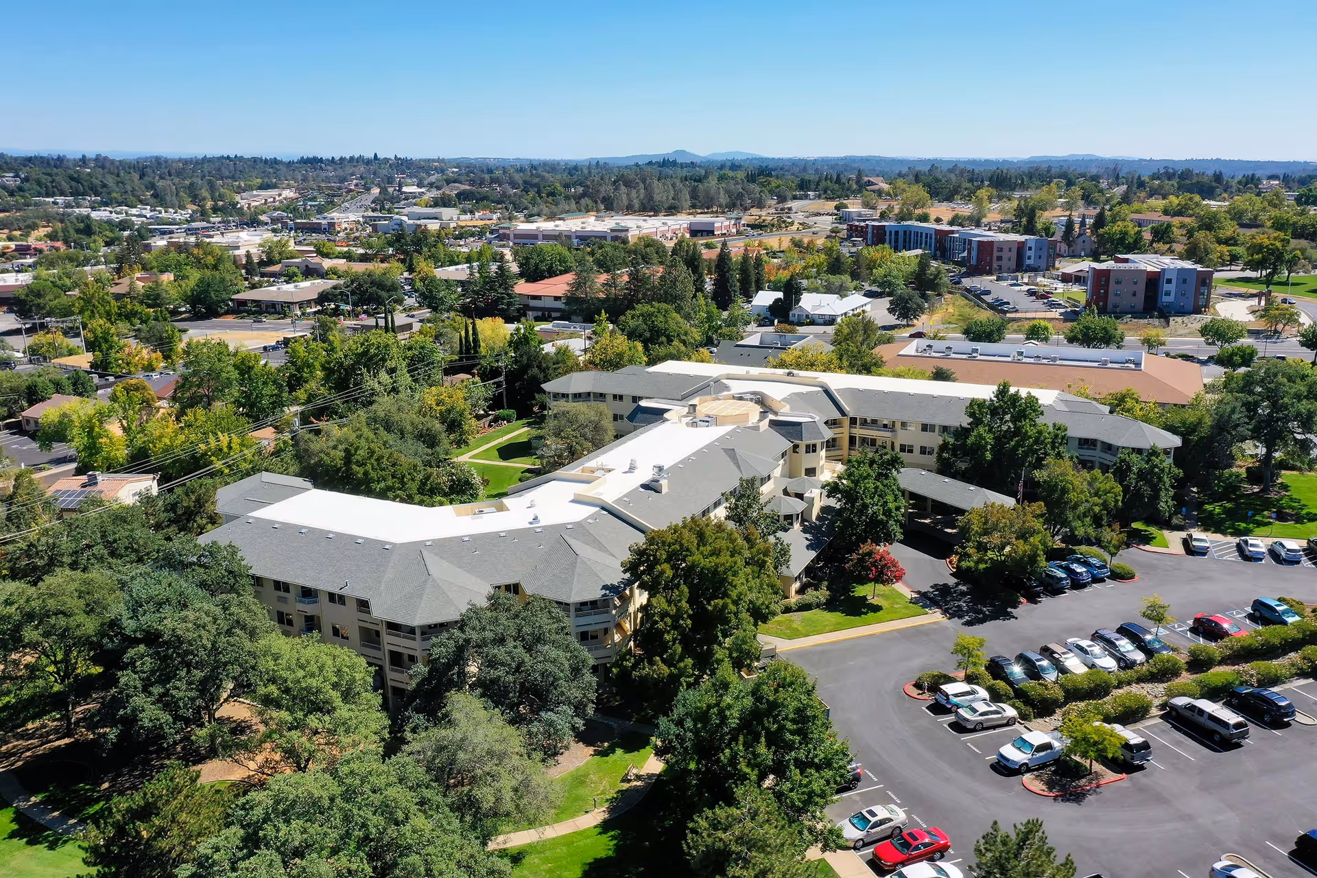 Aerial view of Solstice Senior Living At Auburn, showing a large multi-wing building surrounded by trees and greenery. There is a parking lot with several cars parked, and the surrounding area includes residential and commercial buildings under a clear blue sky.