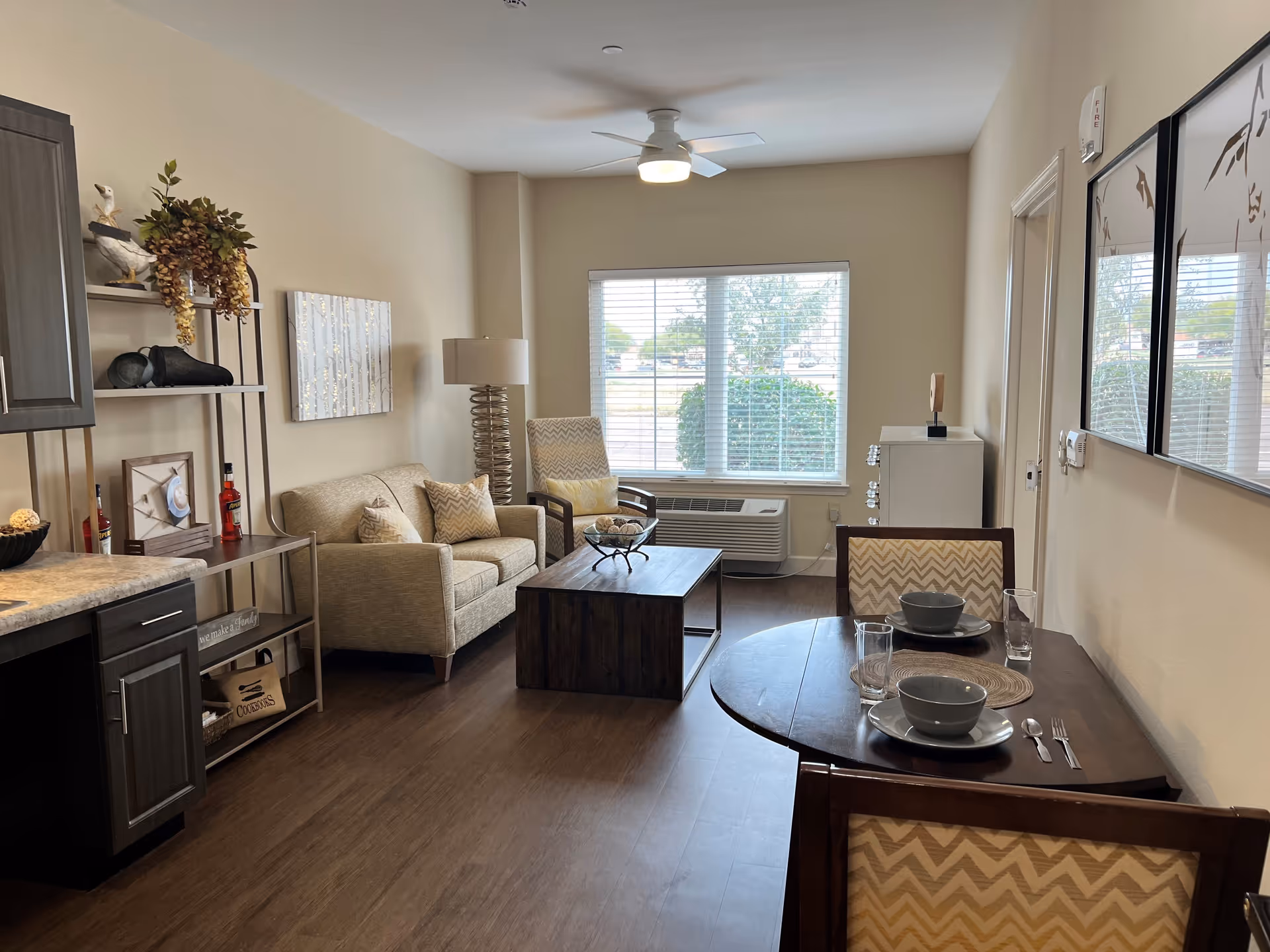 A cozy living room area with a beige sofa, an armchair, a wooden coffee table, and a small dining table set for two with bowls, plates, glasses, and cutlery. The room has a large window with blinds, a ceiling fan with light, and decorative wall art. There is also a shelving unit with decorative items and a lamp with a spiral base.